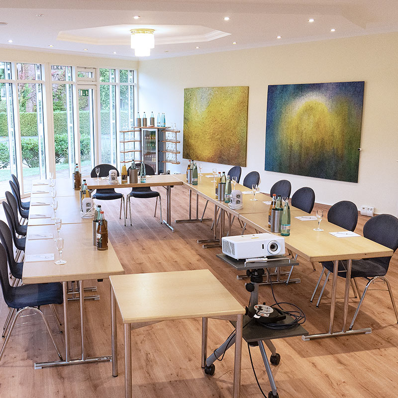 Tagungsraum Lütjenburg Tagungsraum Lütjenburg im Hotel Hohe Wacht, Tische in Stühle in U-Form aufgestellt, eingedeckt mit Gläsern, Getränken und Schreibblöcken, im Vordergrund Beamer, Kronleuchter an Decke, Fensterfront, an der rechten Wand hängen zwei große gemalte Bilder
