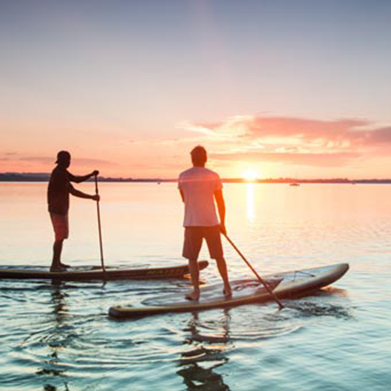 Teambuilding Incentives zwei Personen in kurzen Hosen beim Stand-up-Paddling, auf dem Meer bei Abendstimmung, im Hintergrund Sonnenuntergang