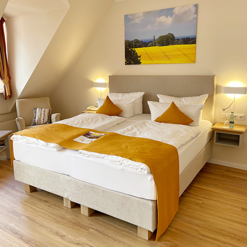 Doppelzimmer Boxspringbett Zimmer mit Doppelbett, am Kopfende Leinwand mit Rapsmotiv, daneben Lampe an der Wand, links vom Bett am Fenster kleine Sitzecke mit Sessel und Tisch