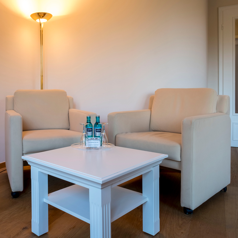 1-Zimmer-Komfortappartement Sitzbereich 1-Zimmer-Komfortappartement Sitzbereich, zwei Sessel mit einem Tisch, hinter dem Sessel steht eine Stehlampe, auf dem Tisch