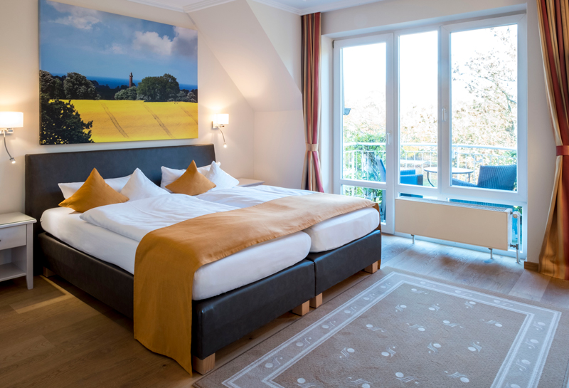 1-Zimmer-Komfortappartement Schlafbereich Schlafbereich im 1-Zimmer-Komfortappartement mit Schlafzimmer, an der Wand Leinwand mit Rapsmotiv, links vom Bett Fenster mit Terrassentür zum Balkon mit Sitzmöglichkeiten und kleinem Tisch, vor dem Bett liegt ein Teppich