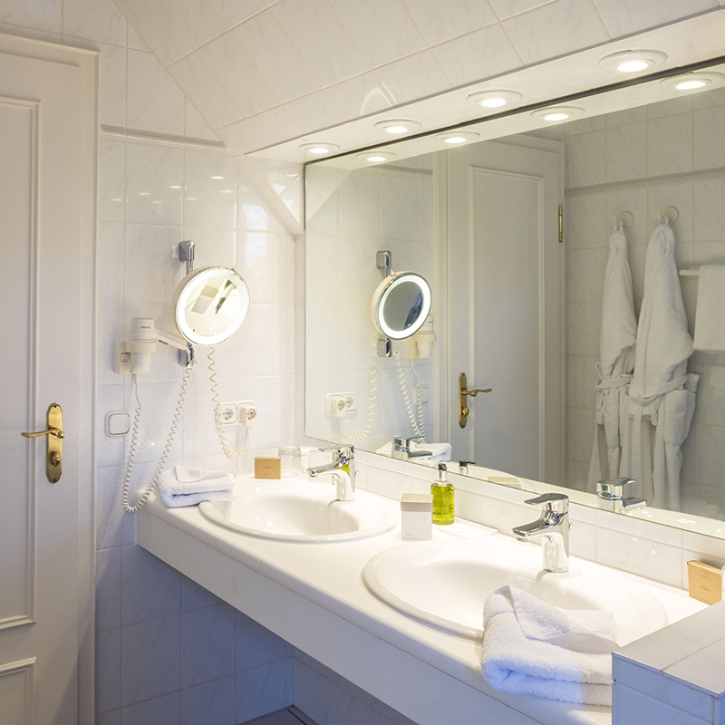 2-Zimmer-Komfortappartement Bad 2-Zimmer-Komfortappartement Bad mit zwei Waschbecken, Spiegelfront, Kosmetikspiegel, Föhn, Handtücher, Kosmetikproben, im Spiegel spiegeln sich zwei Bademäntel hängend am Haken