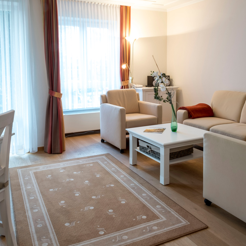 2-Zimmer-Komfortappartement Sitzecke 2-Zimmer-Komfortappartement mit Sitzecke am Fenster, Sofa mit roter Decke, zwei Sessel und Tisch, in der Ecke Sideboard mit Radio und Boxen