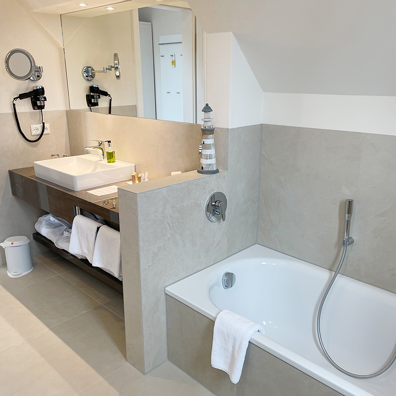 Ostsee-Suite Bad Waschbecken mit Seifenspender, Kosmetikproben, Föhn, Kosmetikspiegel, Handtücher und Kosmetikeimer, Spiegelfront, hinter einer Schamwand Badewanne, über dem Wannenrand hängt ein Handtuch