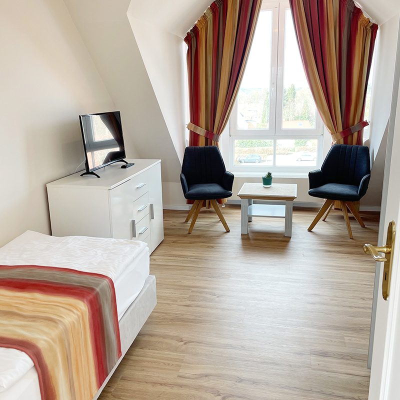 Kurpark-Suite Schlafzimmer Schlafzimmer in Kurpark-Suite, links an der Wand Bett und Sideboard mit TV-Gerät, geradeaus durch Fenster, vor dem Fenster Sitzecke mit zwei Stühlen und Tisch