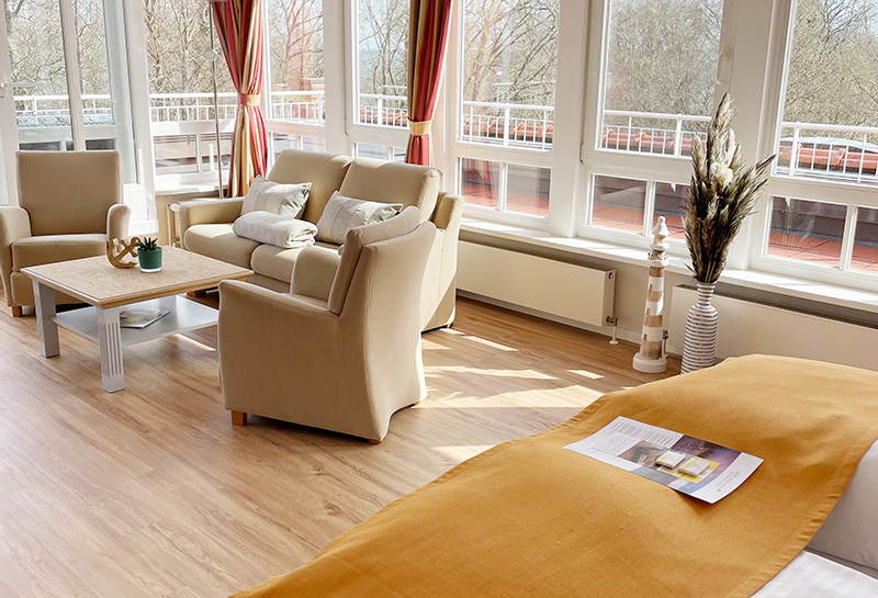 Ostseesuite Sitzecke Suite mit Wohn- und Schlafbereich, Sitzecke mit Sofa, Sessel und Tisch, Doppelbett mit Tagesdecke und Prospekt, helle Fensterfront, umliegender Balkon mit Blick auf die Ostsee