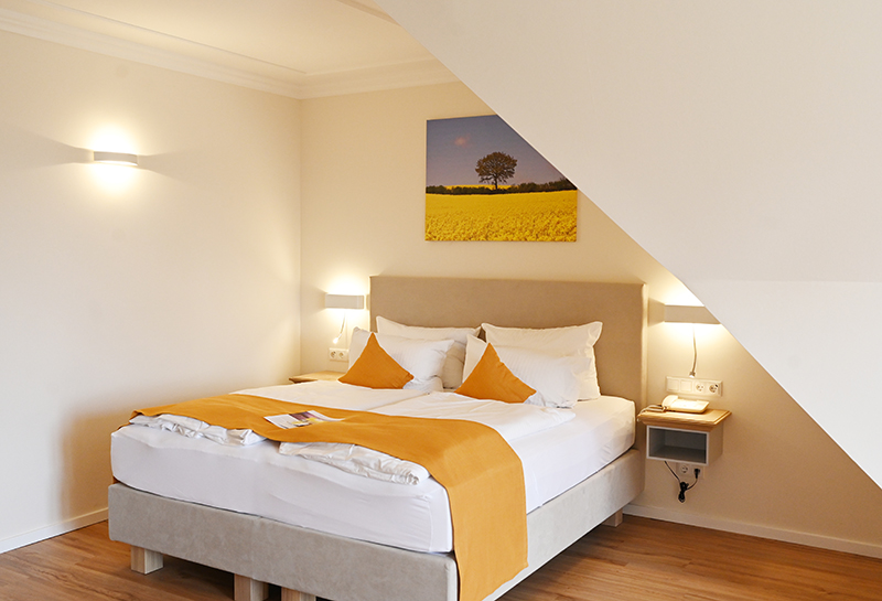 Turmzimmer Schlafbereich Boxspringbett unter der Schräge, über dem Kopfende Leinwand mit Rapsmotiv, links und rechts vom Bett Lampen und Ablageflächen